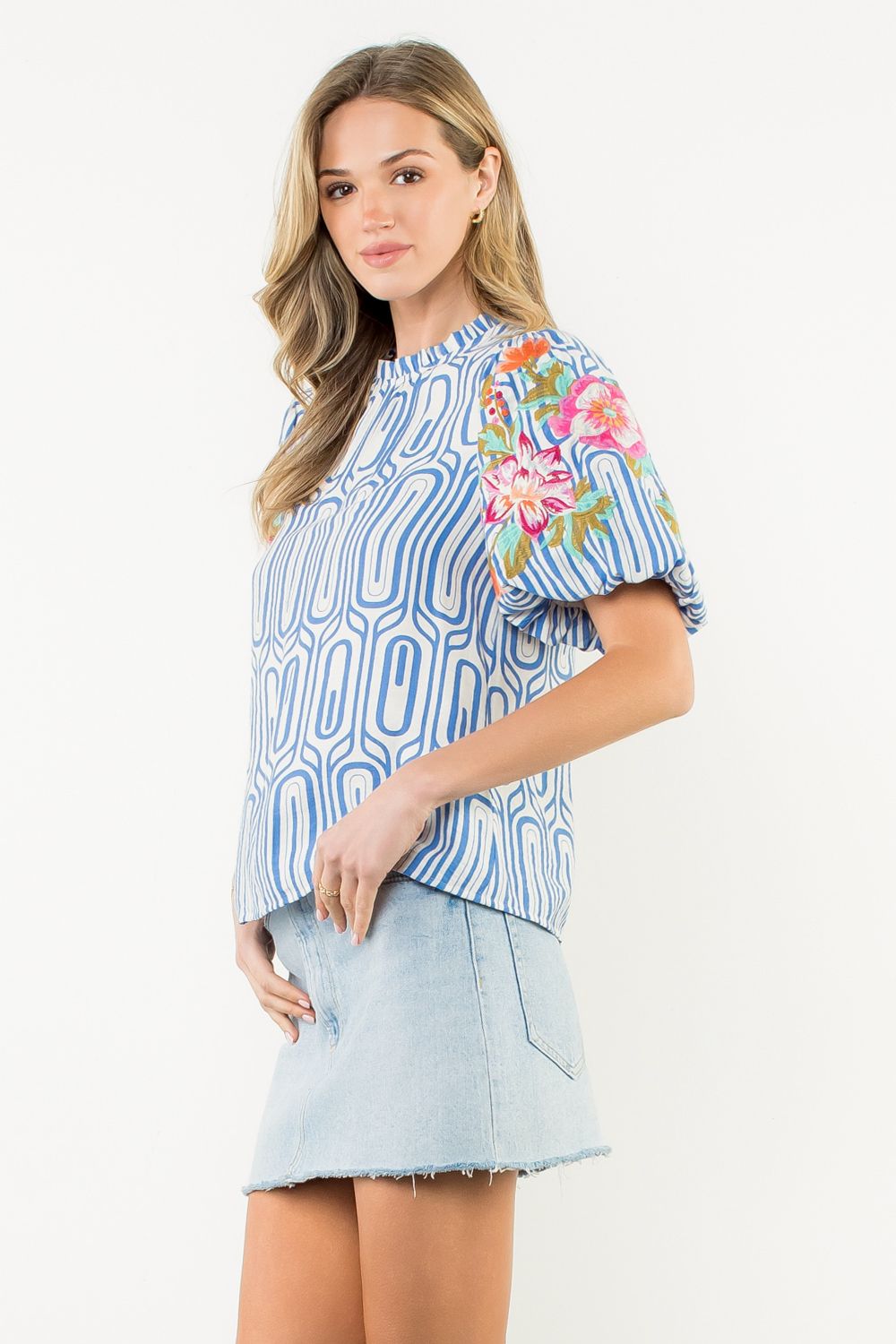 Woman wearing a blue patterned top with floral embroidery and a denim skirt on a white background