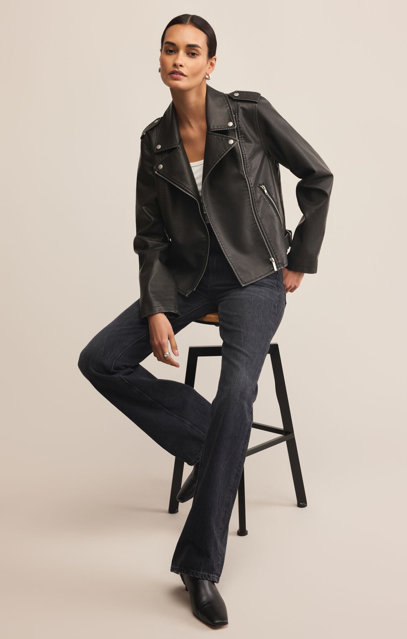 Woman wearing a black leather jacket and dark jeans sitting on a stool against a beige background