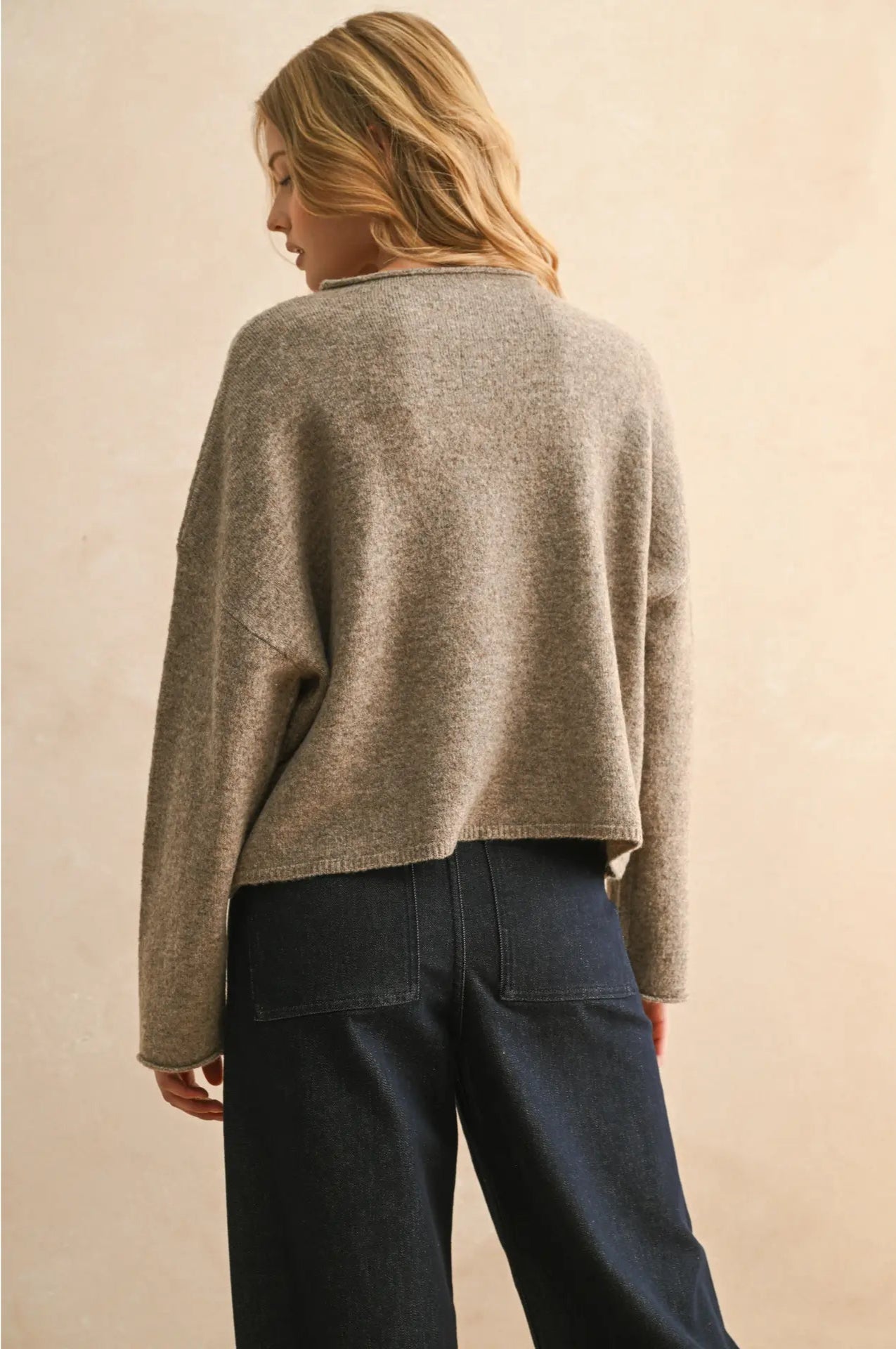 Person wearing a beige sweater and dark jeans against a beige background