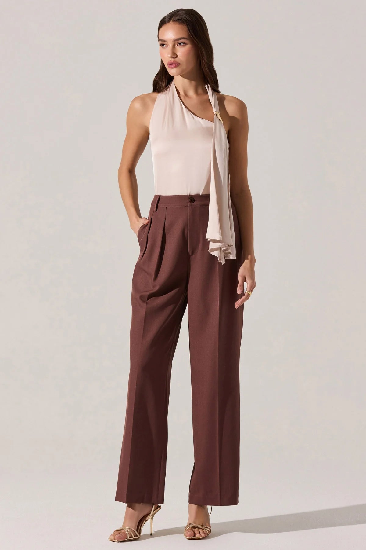 Woman wearing a light pink sleeveless top and brown pants on a plain background