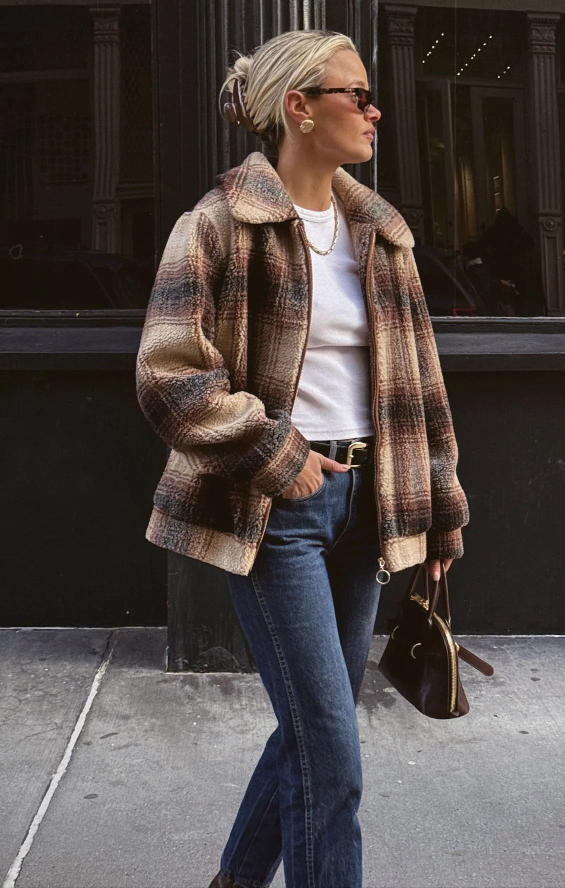 Woman wearing a plaid coat, white shirt, and blue jeans walking on a street.