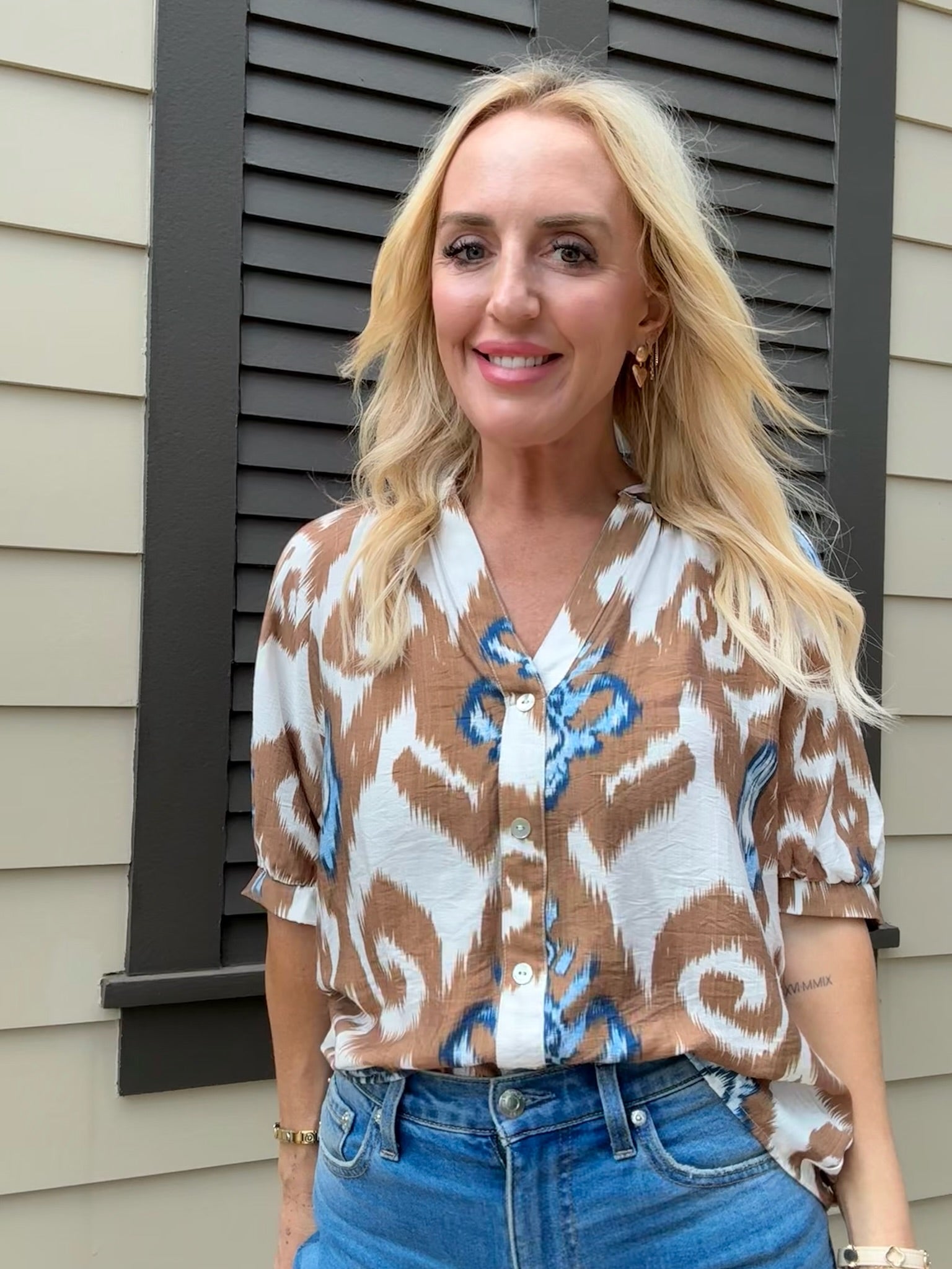 Woman wearing a patterned blouse and blue jeans standing in front of a house.
