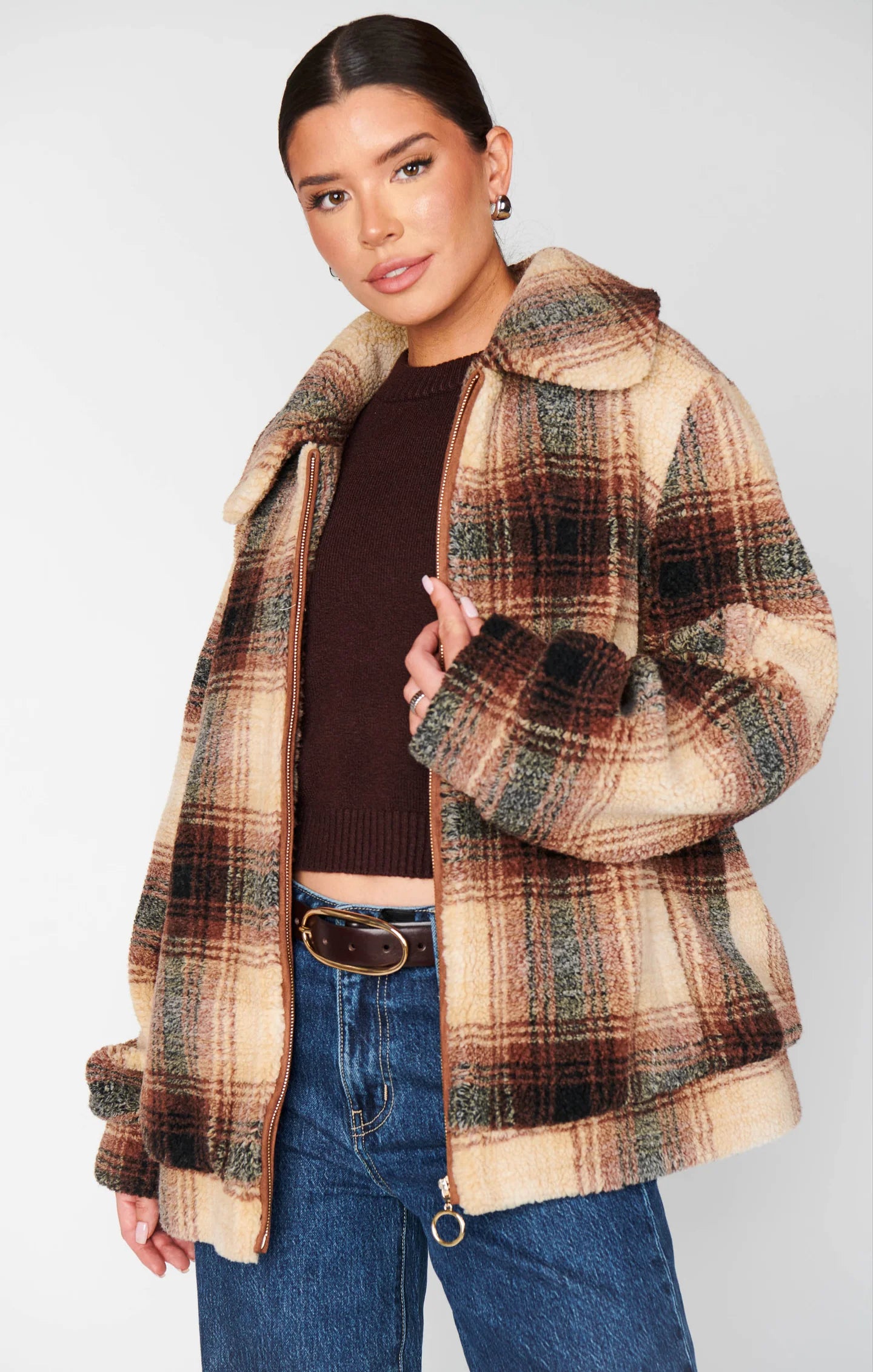 Woman wearing a plaid coat over a brown sweater and blue jeans on a white background