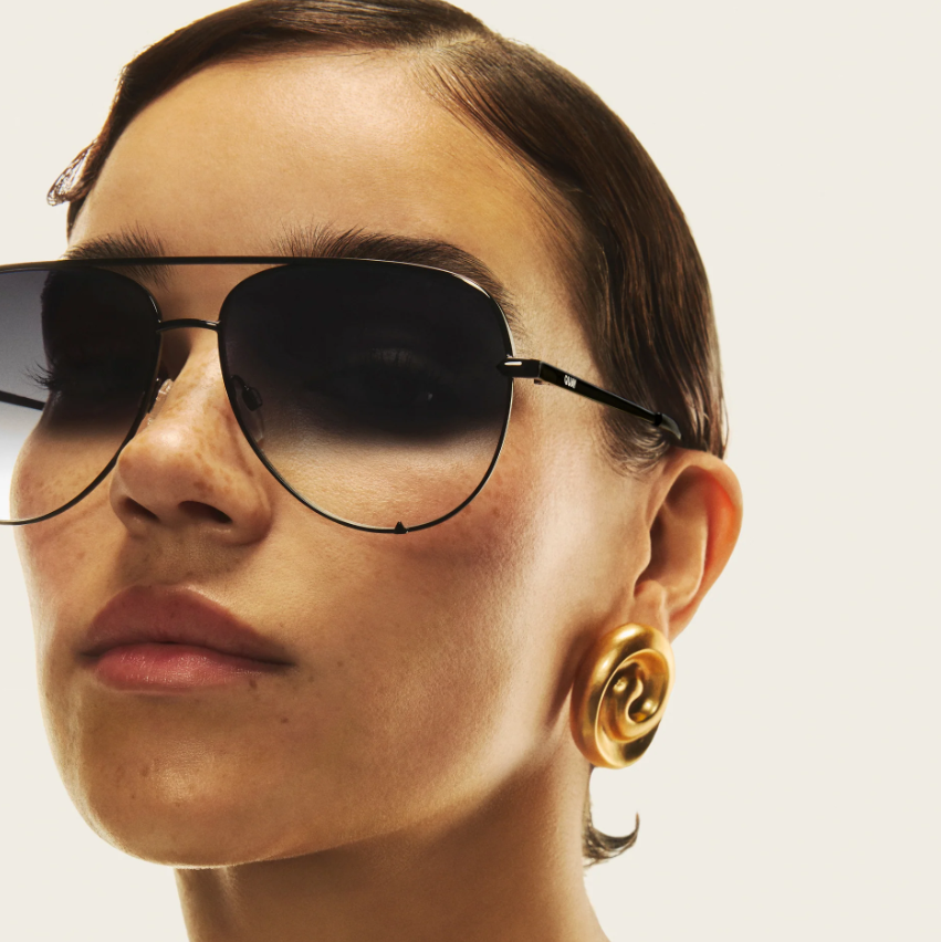 Person wearing sunglasses and gold earrings on a neutral background