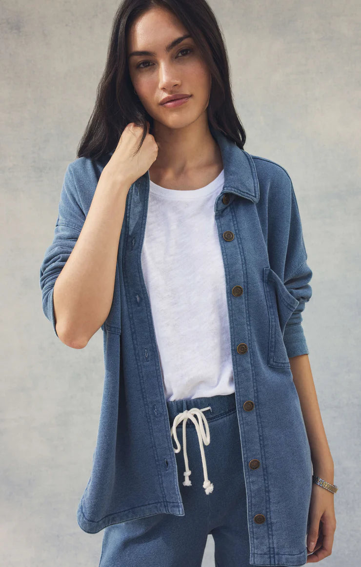 Woman wearing a denim jacket over a white shirt and blue pants against a plain background