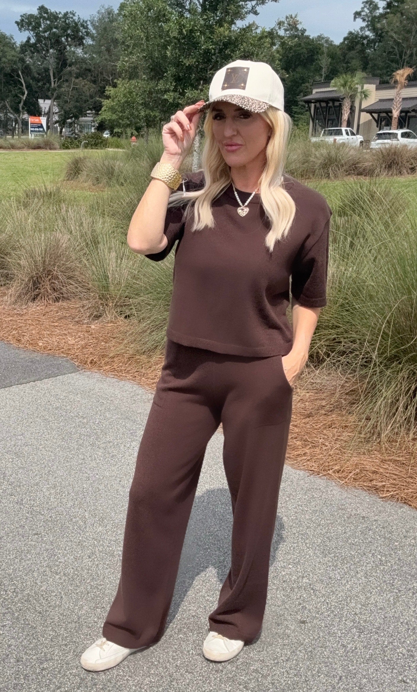 Woman in brown outfit and cap standing outdoors on a path with greenery.