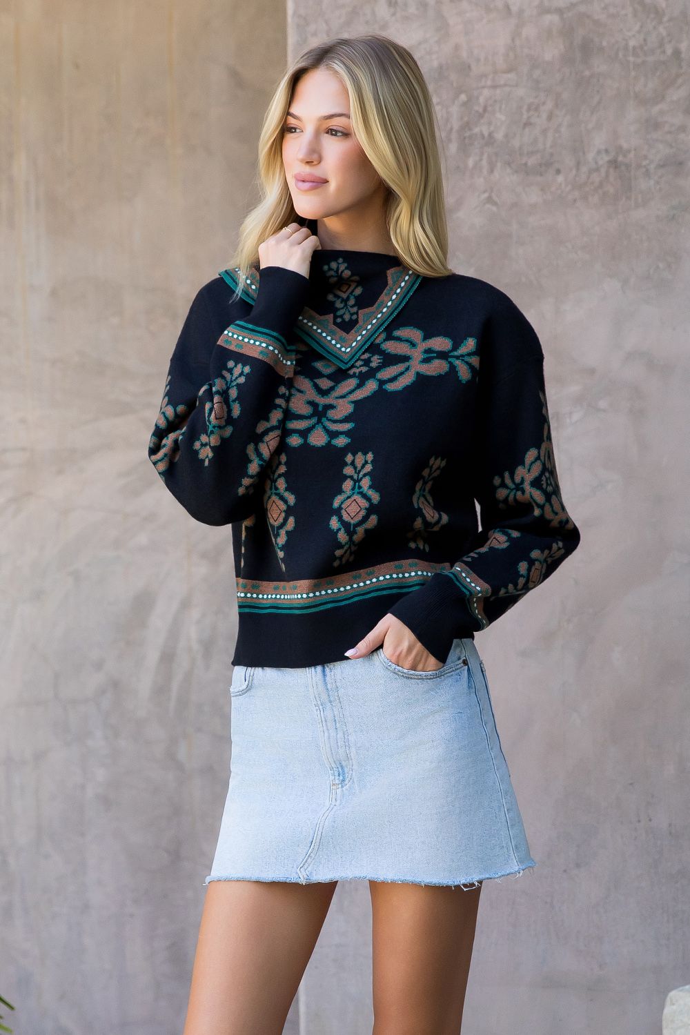 Woman wearing a dark sweater with a pattern and light blue denim skirt against a neutral background