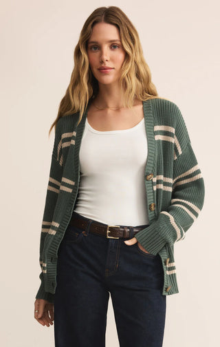 Woman wearing a green cardigan with white stripes, white tank top, and dark pants on a beige background