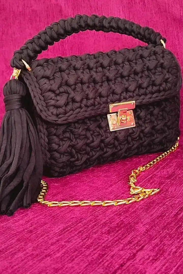 Black textured handbag with a gold chain and tassel on a pink background