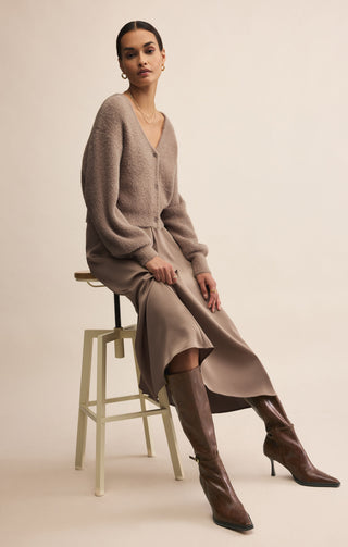 Woman wearing a long beige sweater dress and brown knee-high boots sitting on a stool.