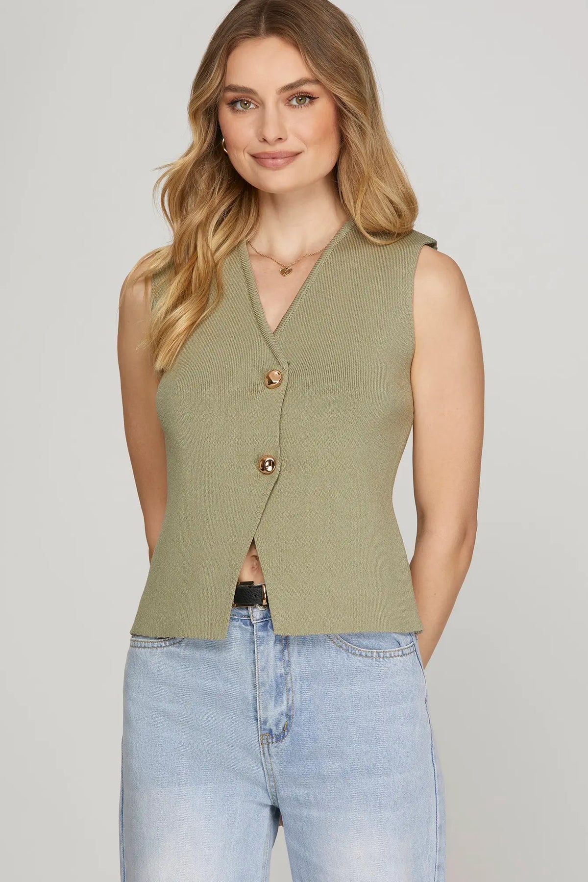 Woman wearing a green vest over a black top and blue jeans on a gray background