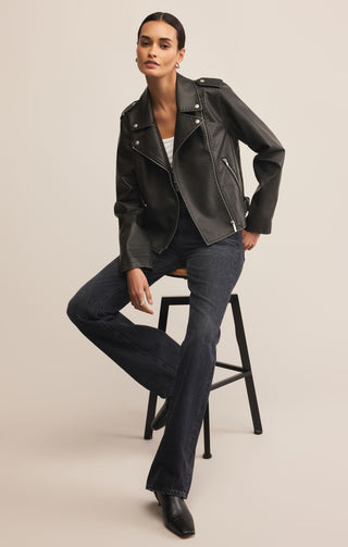 Woman wearing a black leather jacket and dark jeans sitting on a stool against a beige background