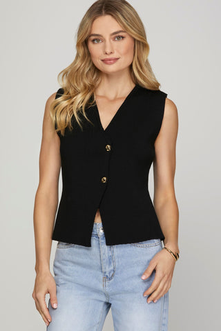 A woman wearing a sleeveless black sweater vest with gold buttons, paired with blue jeans.