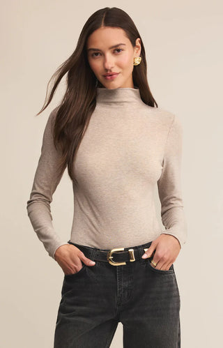 Woman wearing a beige turtleneck sweater and black pants against a plain background