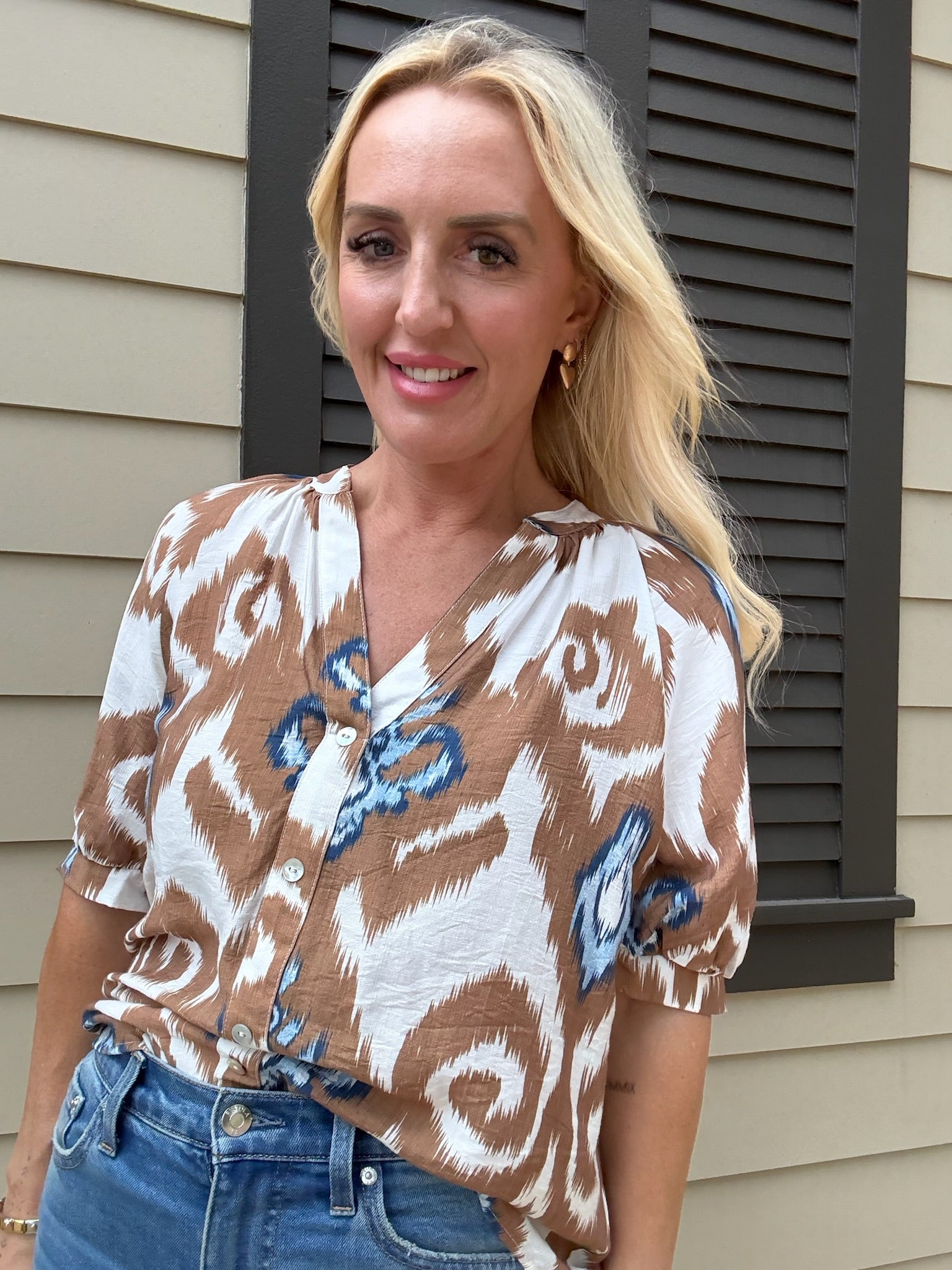 Woman wearing a patterned blouse and blue jeans standing in front of a house.