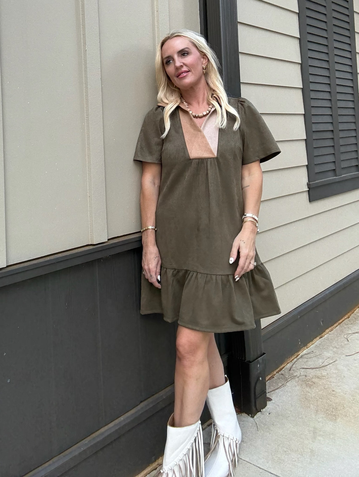 Woman wearing an olive green dress with a sheer top and white fringe boots standing against a house exterior.