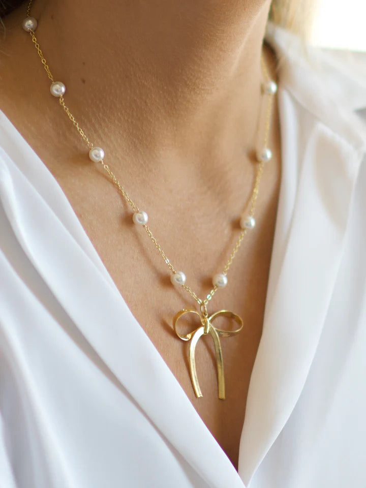 Gold necklace with a bow pendant and pearl beads worn by a person in a white outfit.