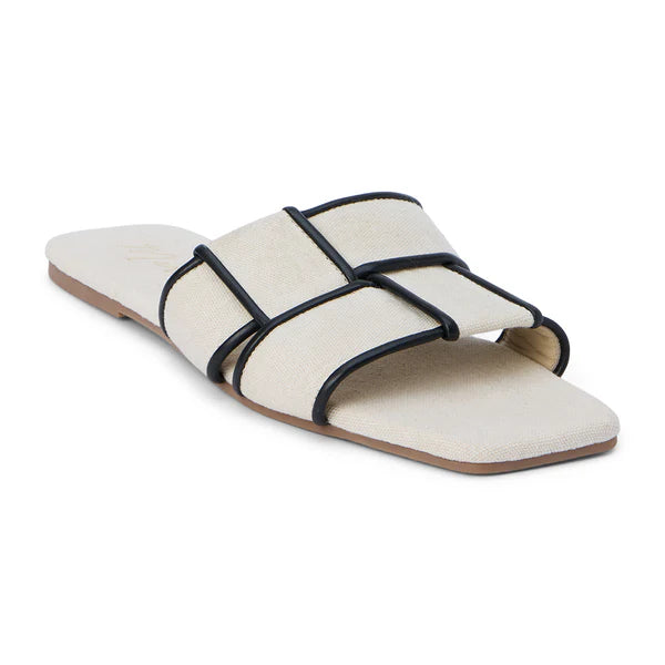 A pair of flat slide sandals with woven bands and squared toes, displayed on a wooden surface, likely indoors.
