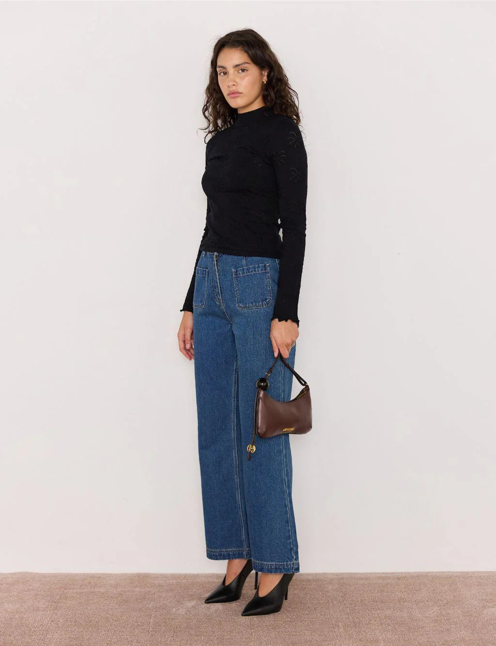 Woman wearing a black top and blue jeans holding a brown handbag against a white background