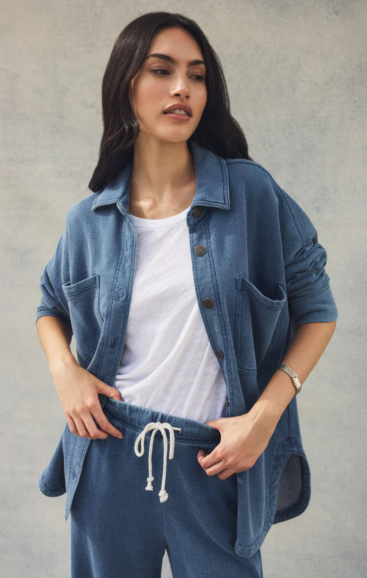 Woman wearing a denim jacket over a white shirt and blue pants against a plain background