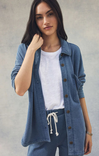 Woman wearing a denim jacket over a white shirt and blue pants against a plain background