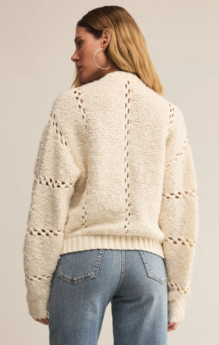 Woman wearing a cream sweater with a textured pattern and blue jeans on a plain background