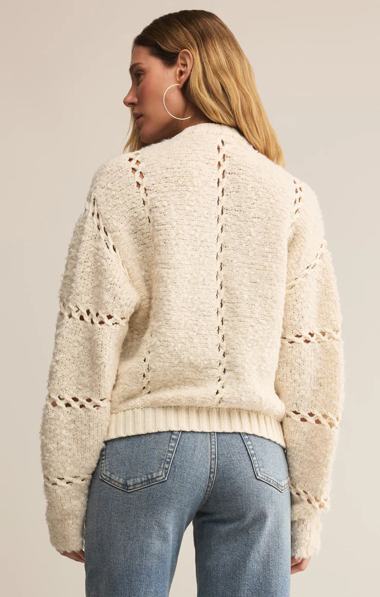 Woman wearing a cream sweater with a textured pattern and blue jeans on a plain background
