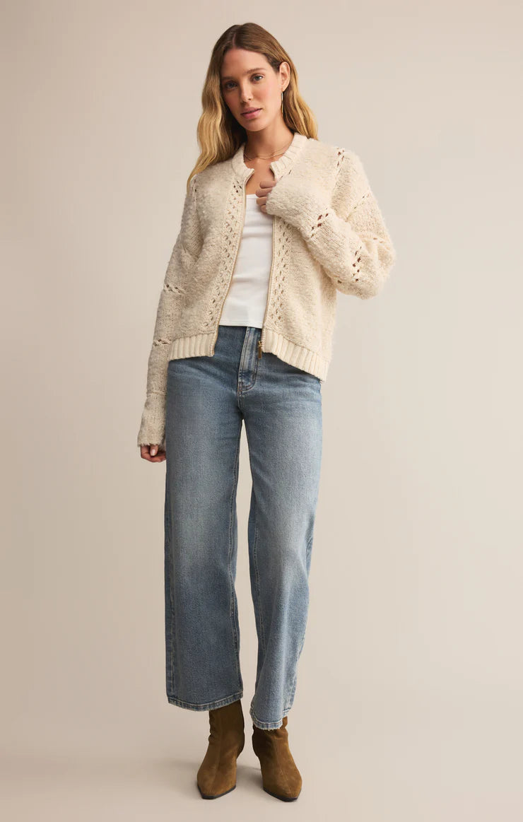 Woman wearing a cream cardigan, white shirt, blue jeans, and brown boots on a beige background