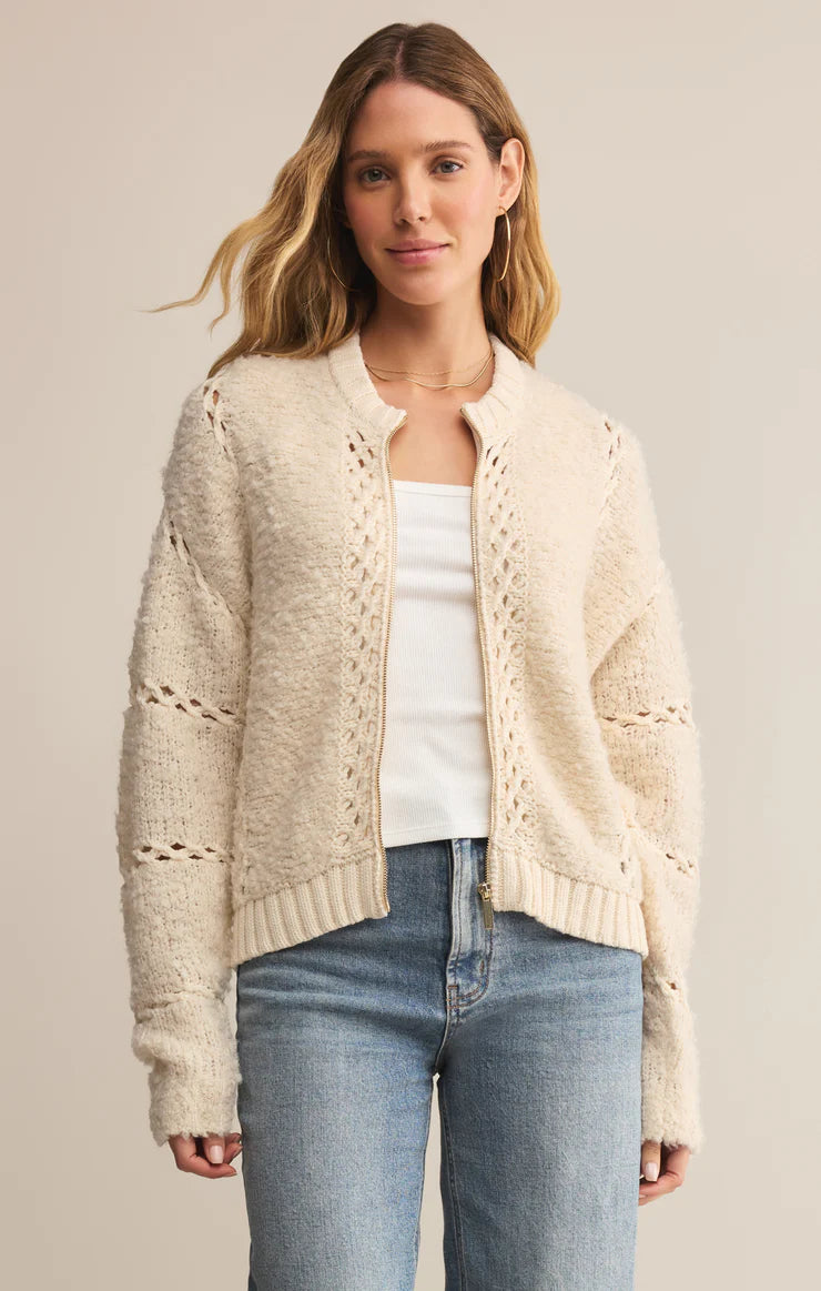 Woman wearing a cream knitted cardigan over a white top and blue jeans on a beige background
