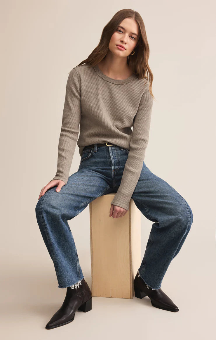 Woman sitting on a cardboard box wearing a gray sweater, blue jeans, and black shoes against a beige background