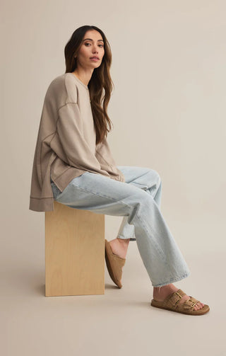 Woman sitting on a wooden block wearing a beige sweater, light blue jeans, and brown sandals against a beige background.