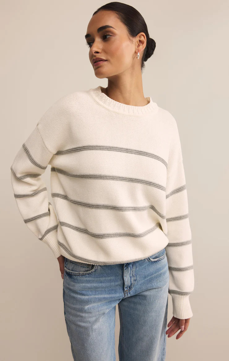 Woman wearing a cream sweater with gray stripes and blue jeans against a plain background