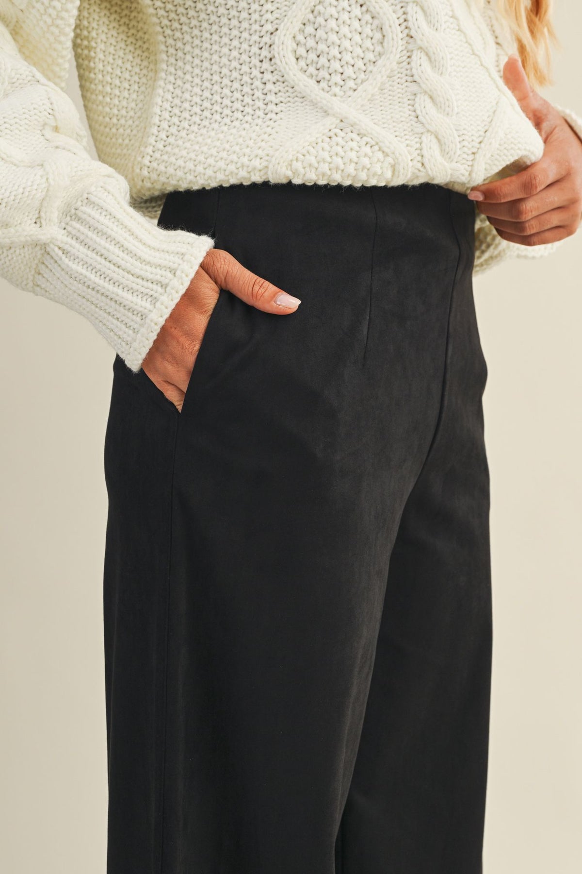 Person wearing a white cable knit sweater and black pants against a neutral background