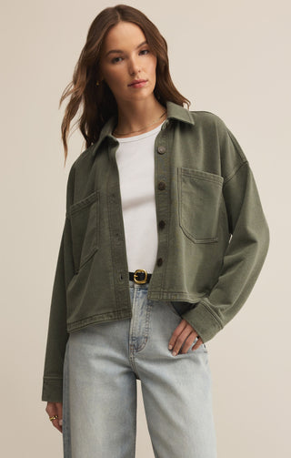 Woman wearing an olive green jacket, white shirt, and light blue jeans on a beige background