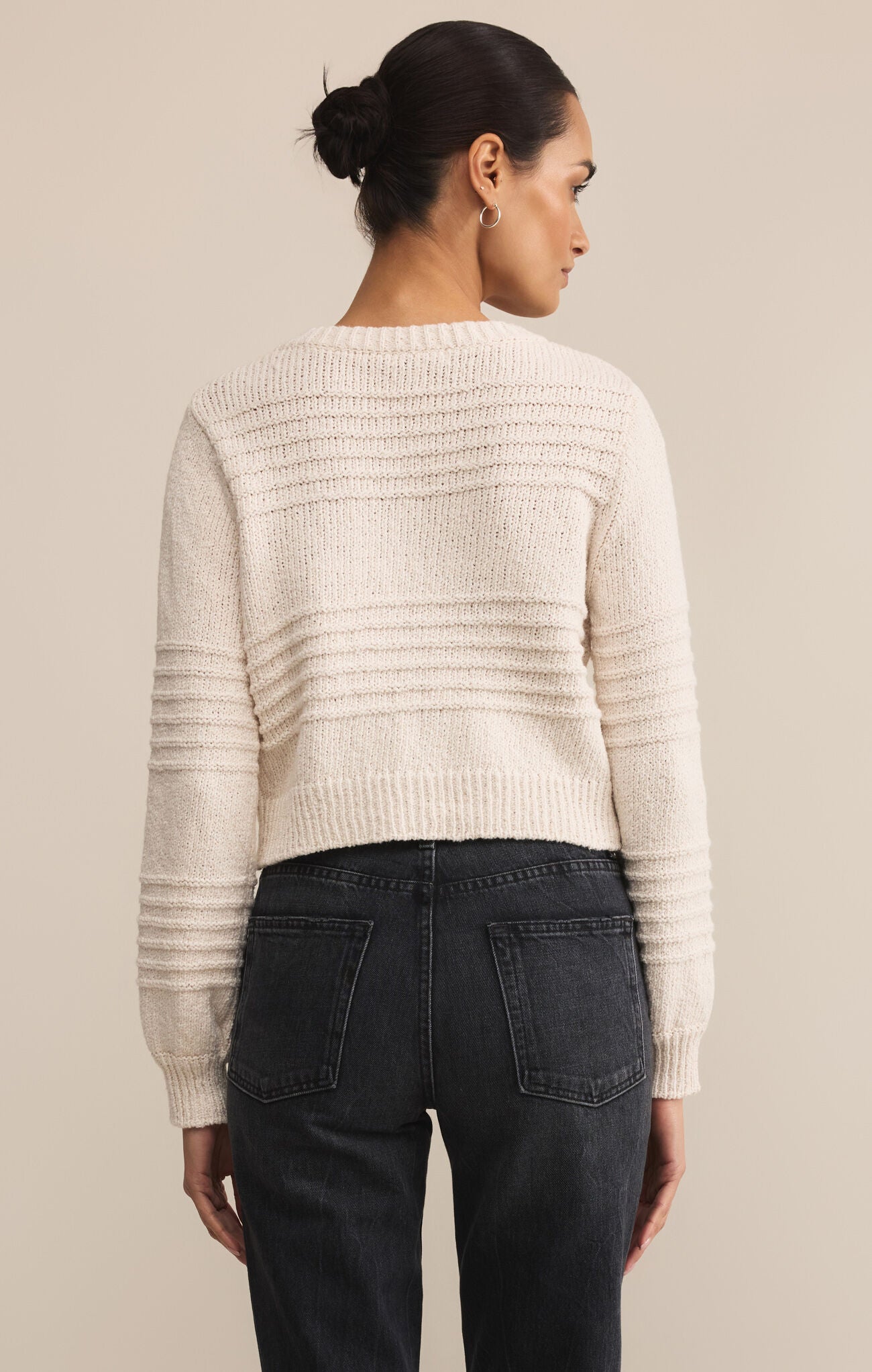 Woman wearing a beige knitted sweater and dark jeans against a beige background
