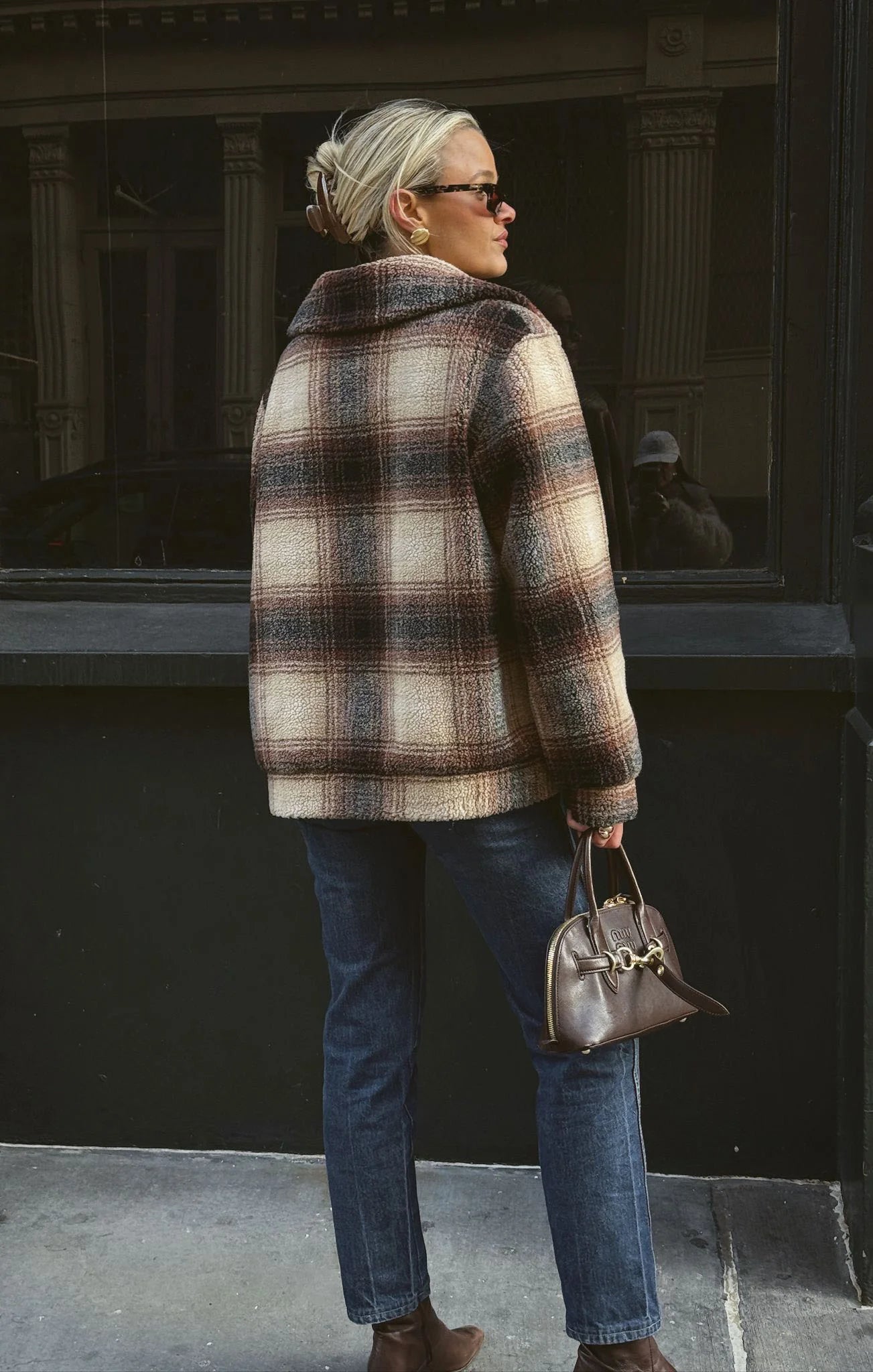 Woman wearing a plaid coat and sunglasses, standing on a street.