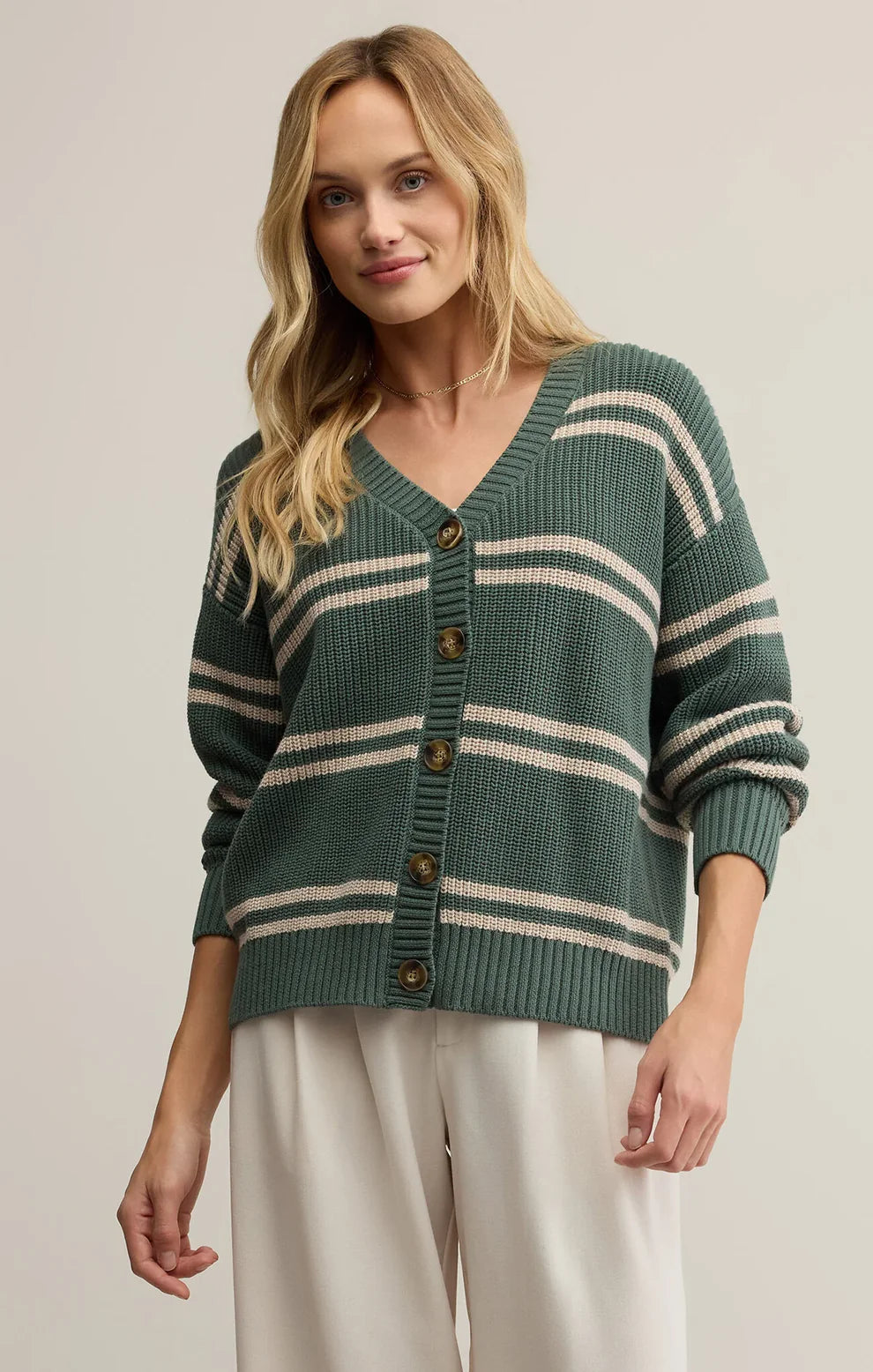 Woman wearing a green cardigan with white stripes, white tank top, and dark pants on a beige background