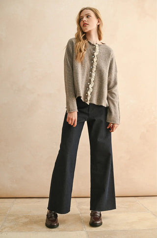 Woman wearing a gray cardigan with ruffled details and black pants against a beige background