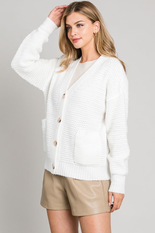 Woman wearing a white cardigan over a beige top and shorts on a plain background