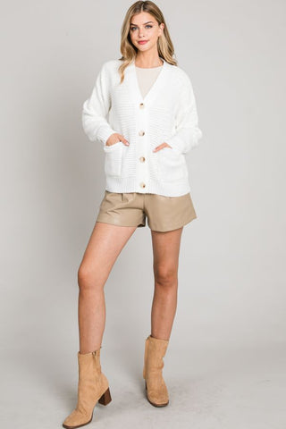 Woman wearing a white cardigan, beige shorts, and tan boots on a plain background