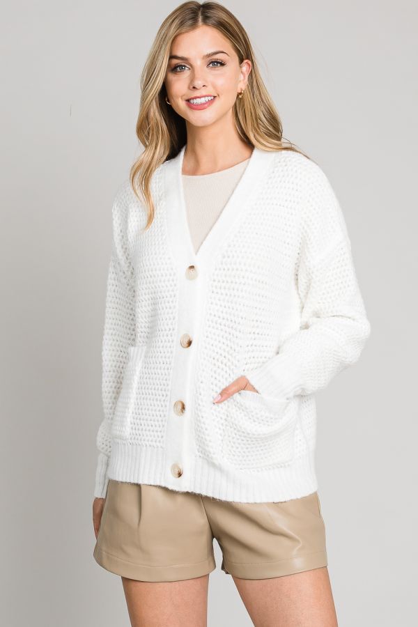 Woman wearing a white cardigan over a beige top and shorts on a plain background