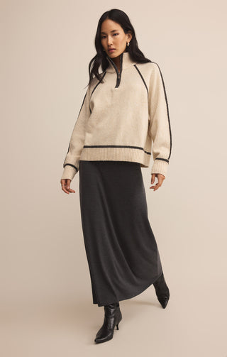 Woman wearing a beige sweater with black trim and a long black skirt on a beige background