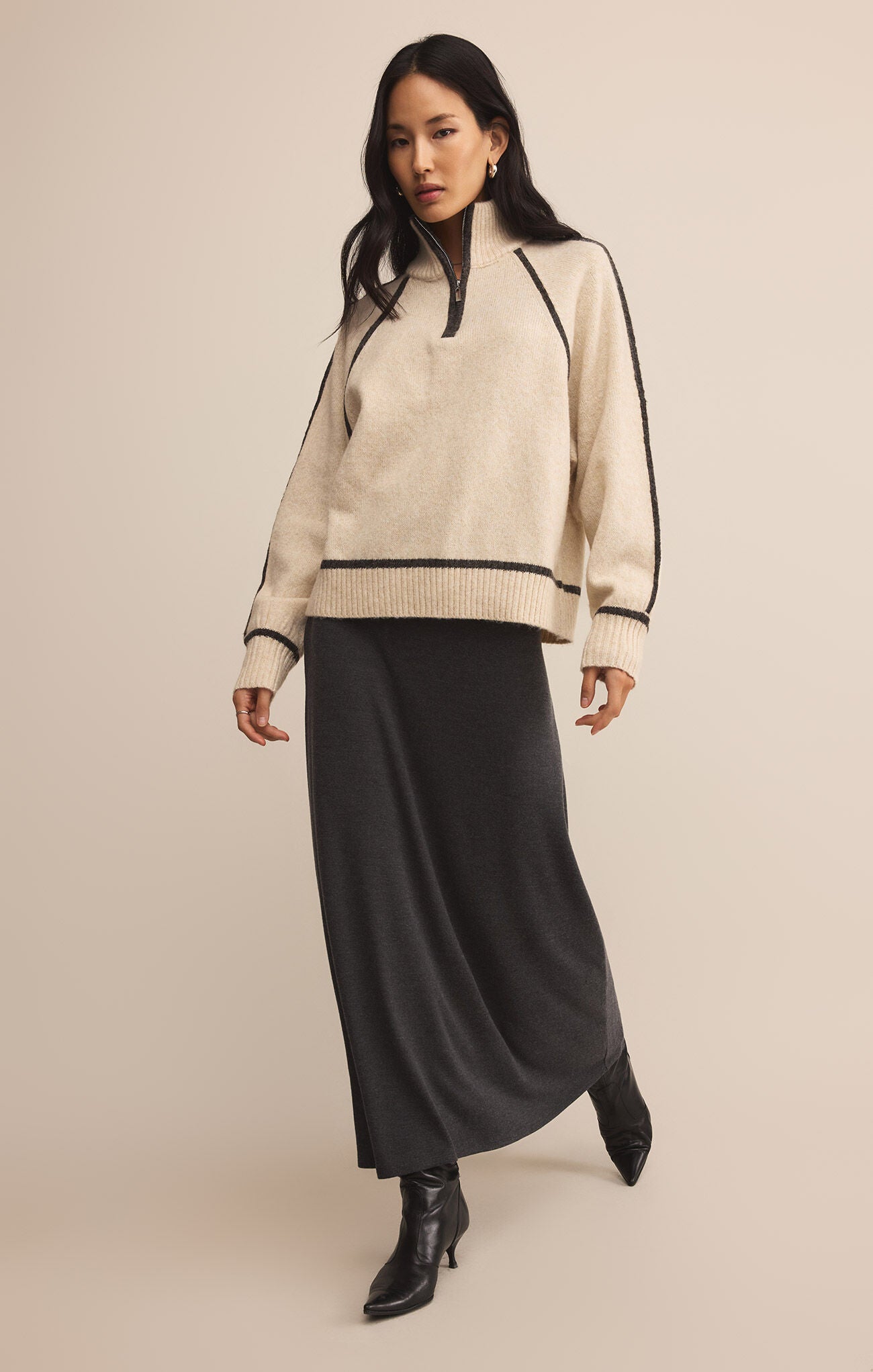 Woman wearing a beige sweater with black trim and a long black skirt on a beige background