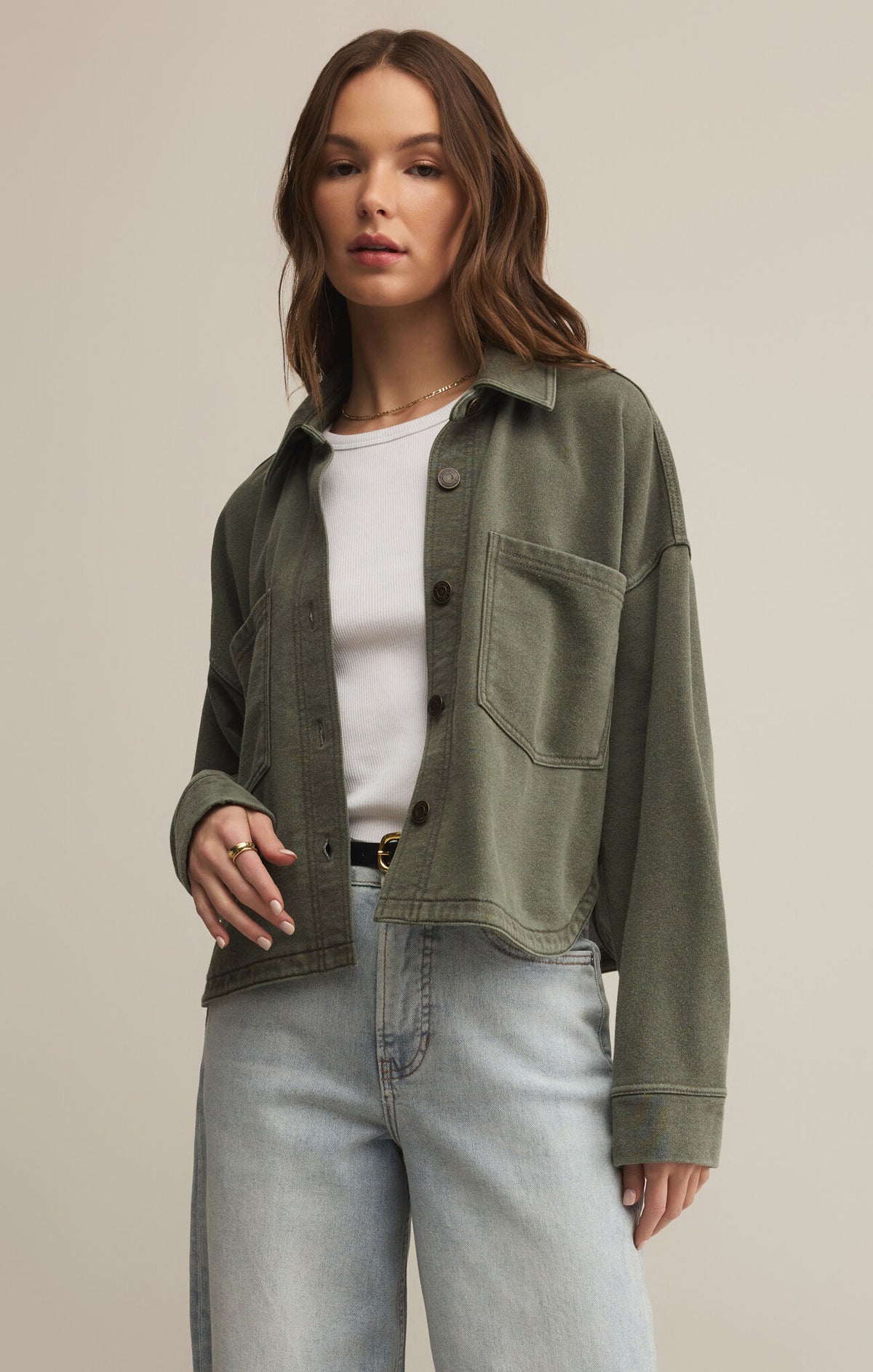 Woman wearing an olive green jacket over a white shirt and light blue jeans on a beige background