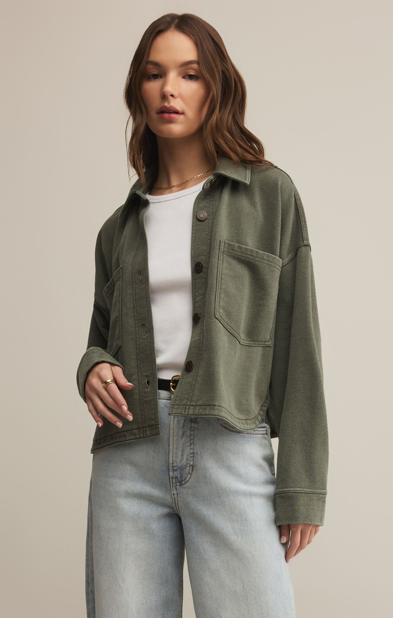 Woman wearing an olive green jacket, white shirt, and light blue jeans on a beige background