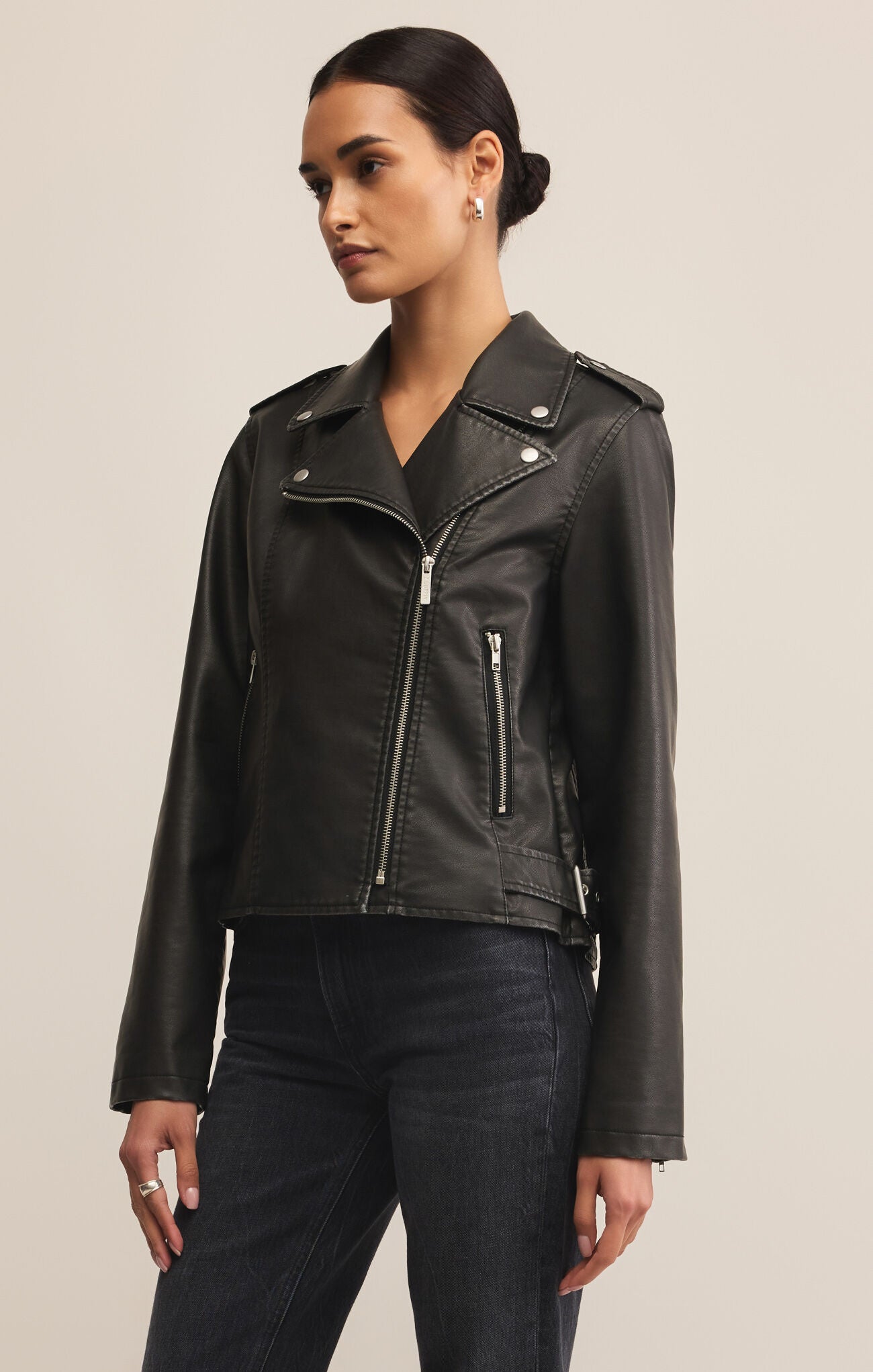 Woman wearing a black leather jacket over a white top and dark jeans on a beige background