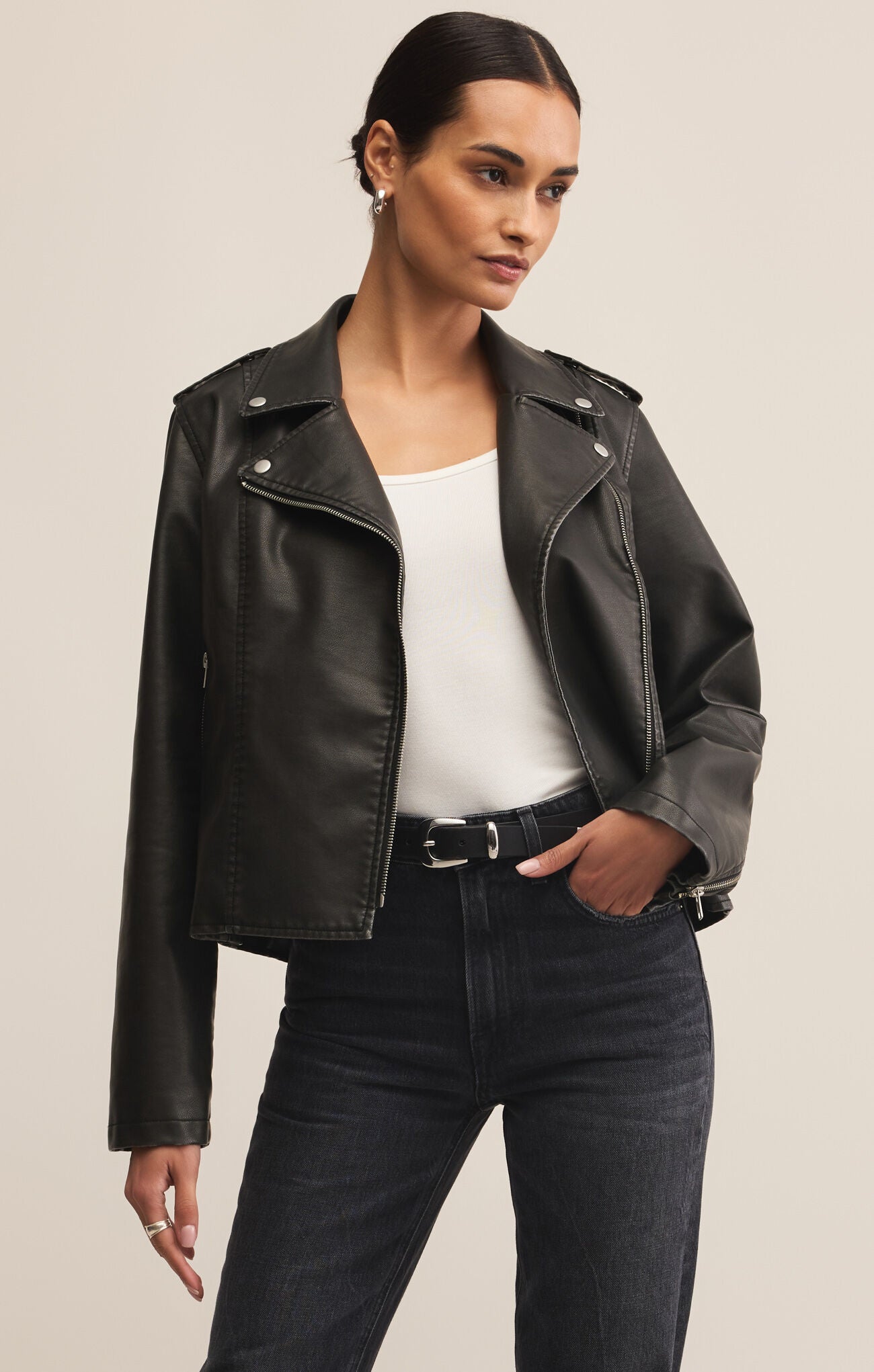 Woman wearing a black leather jacket over a white top and dark jeans on a beige background