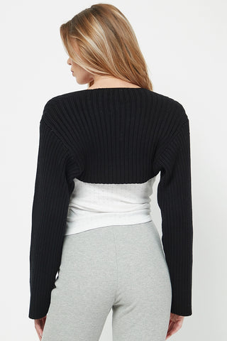 Woman wearing a black ribbed sweater over a white top and gray pants on a white background