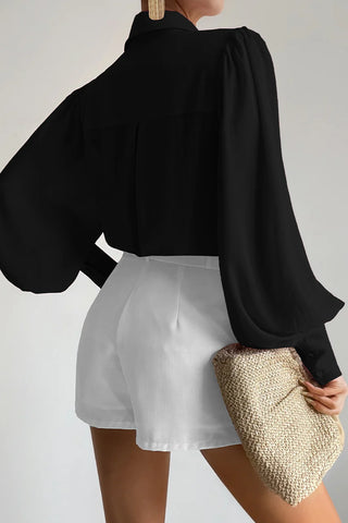 Person wearing a black blouse and white shorts holding a beige clutch against a plain background