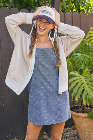 Woman wearing a denim dress and beige cardigan with a white cap, standing outdoors.